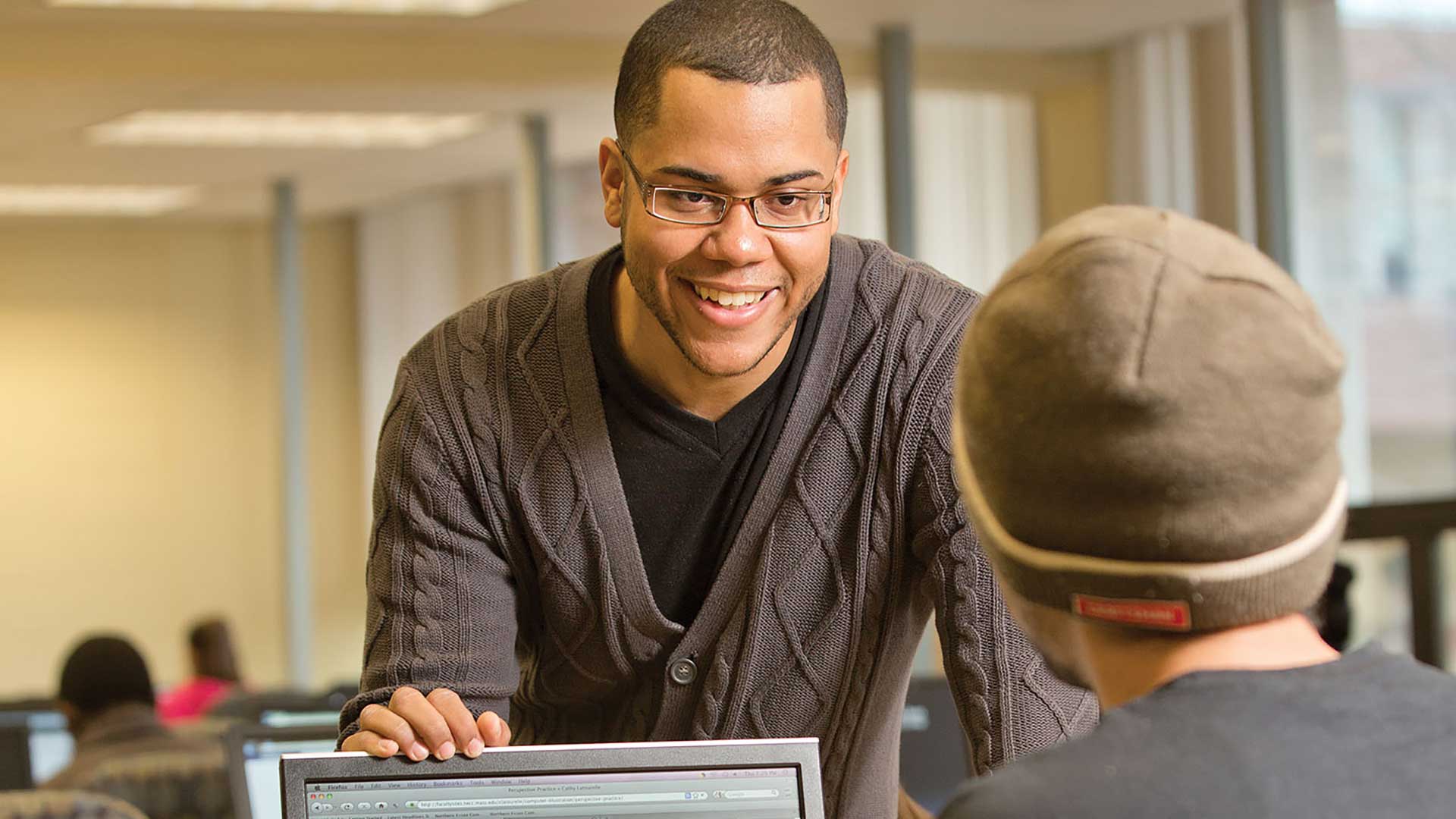 A dude in glasses and sweater is touching the top of another dude's monitor.