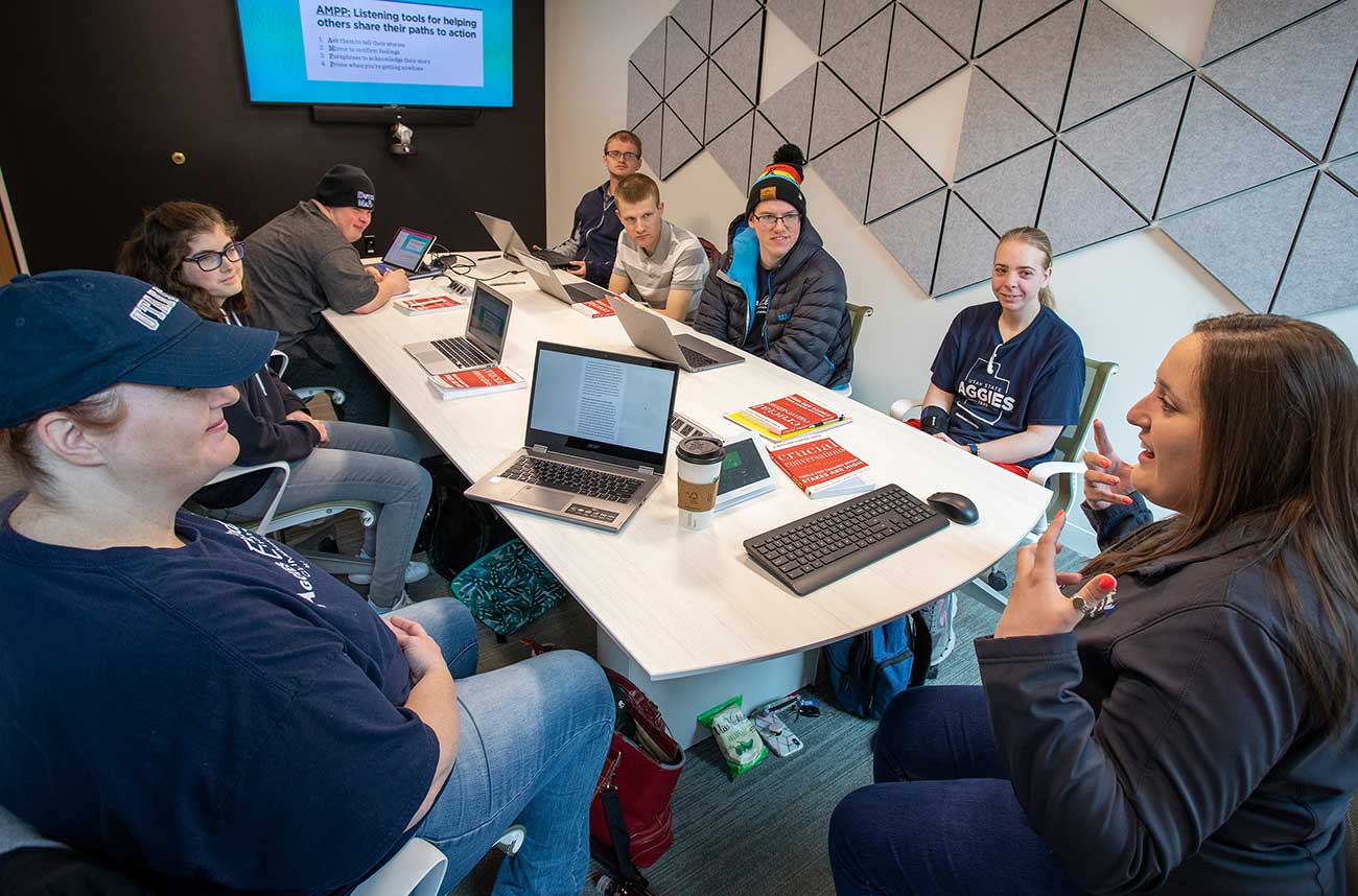 Small conference room with 7 students around a single table with laptops. A wall mounted screen on the far end of the room displays lesson materials.