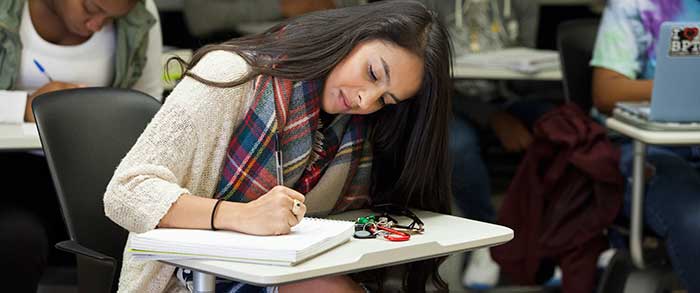 DACA student Laura Piñeros at desk in a full classroom writing in a notebook.