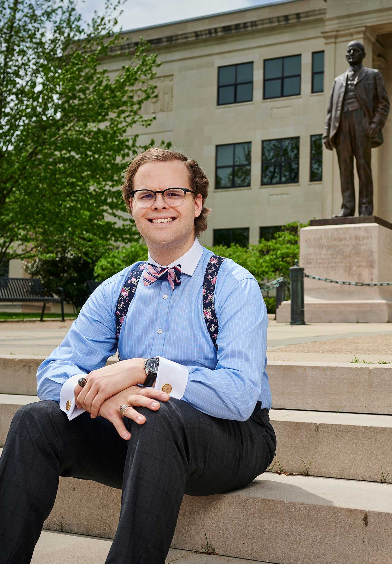 David is well-dressed in this photo with bow-tie and suspenders. He's smiling, sitting on concrete steps of a campus building. An unidentifiable bronze statue is visible in the background.