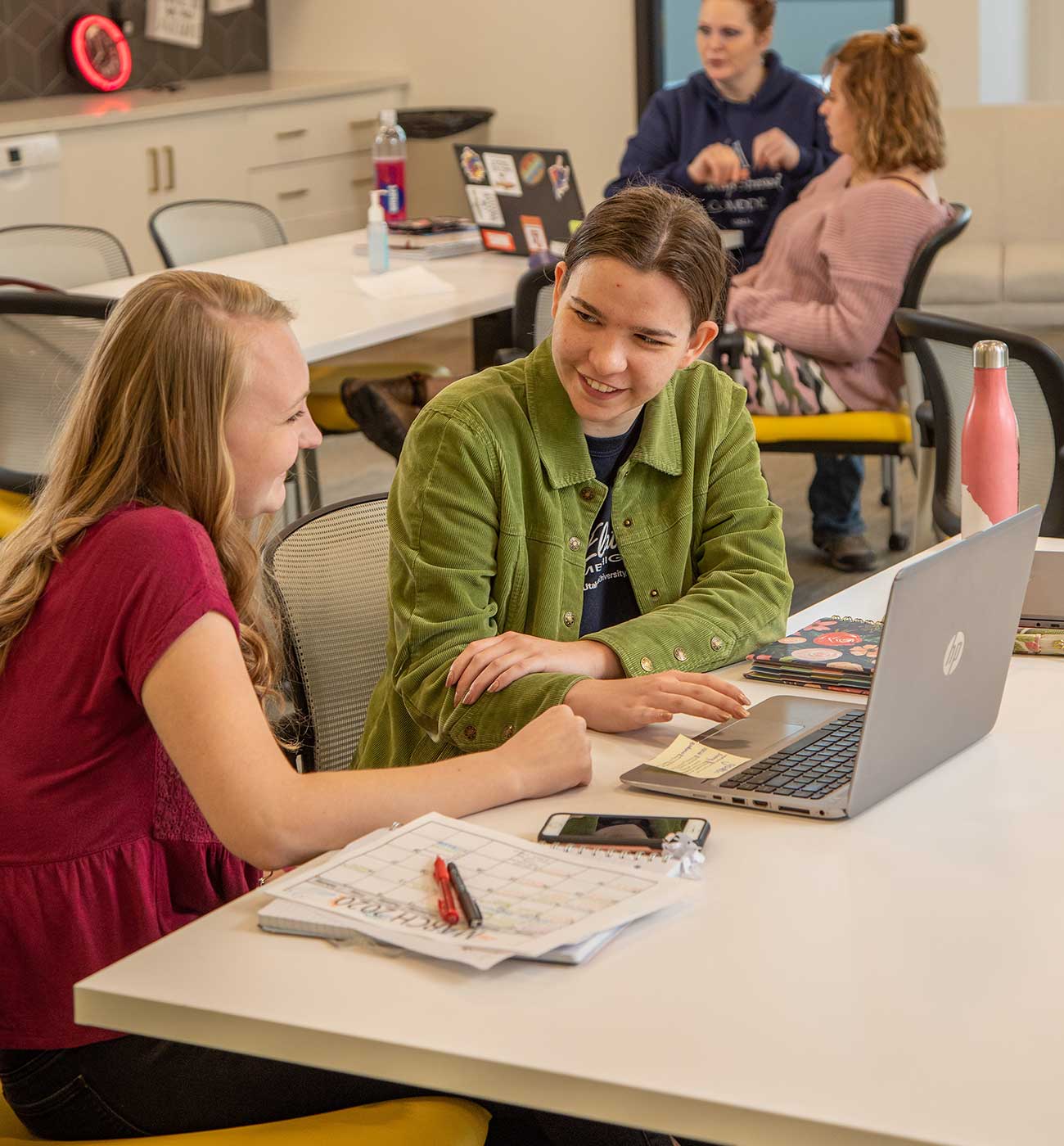 Smiling student and mentor working together at a table. Another pair are seated at another table in the background.