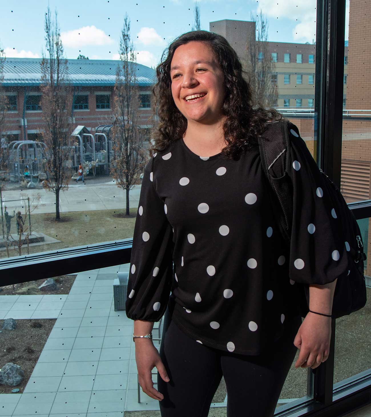 Student Jenna Mosher in polka dot blouse with backpack in a campus building in front of a large multi-paned window. Other buildings and a playground are visible in the background.