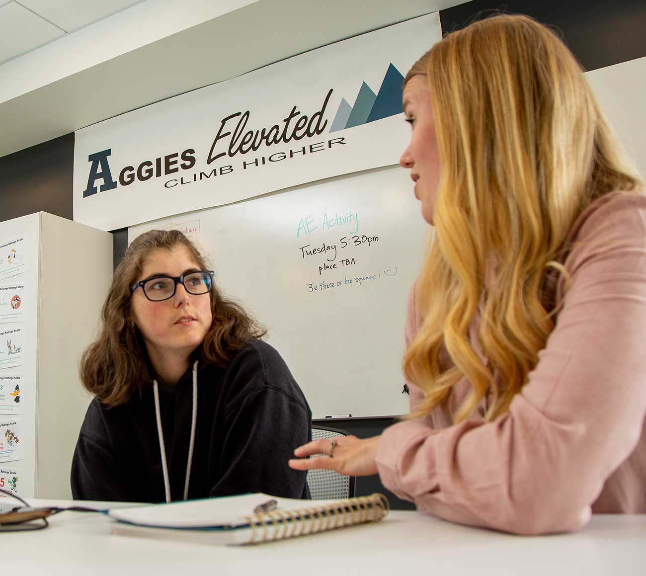 Student and mentor conversing. A spiral bound notebook is on the desk. An Aggies Elevated banner is visible in the background.