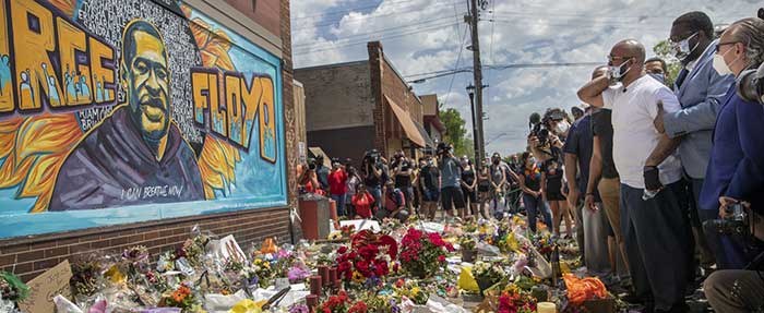 George Floyd memorial mural painted on wall of cup foods where Floyd was arrested. Mourners gather in a circle. The ground is covered with notes, candles, and flowers.