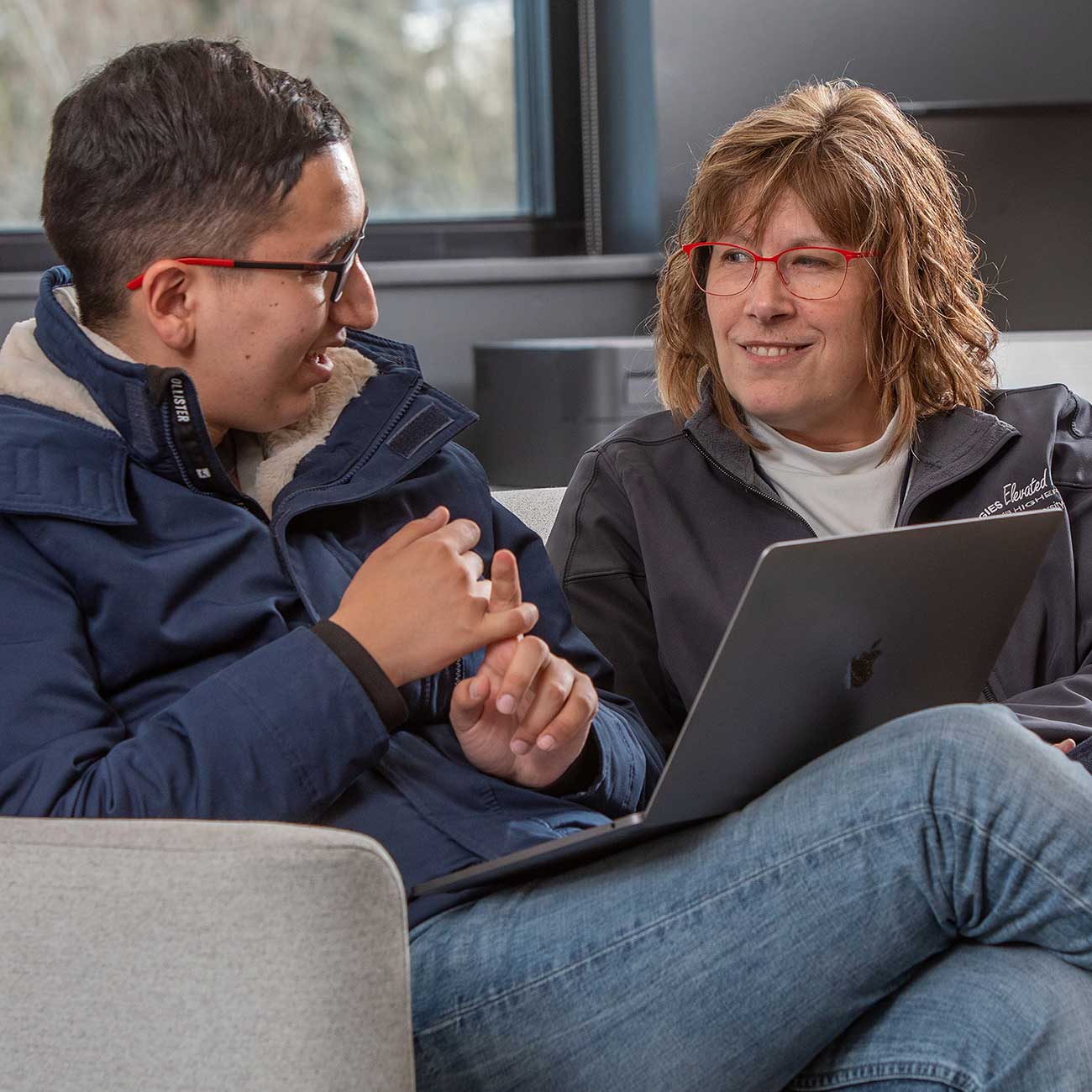 Program director and student with laptop converse on couch in relaxed pose.