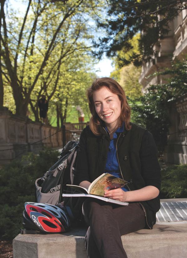 UW graduate Casmir Turnquist-Held is pictured seated on a concrete bench on campus. She has a book and notebook on her lap. Beside her is a large backpack and bike helmet.