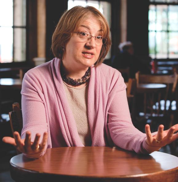 Elaine Klein in pink sweater at a wooden table in a study area gesticulating while talking with her palms upward.