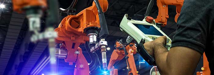 A dark factory floor with 3 large robotic manufacturing arms painted in bright orange. A man at the edge of the photo can be seen controlling the arms with a tablet device.