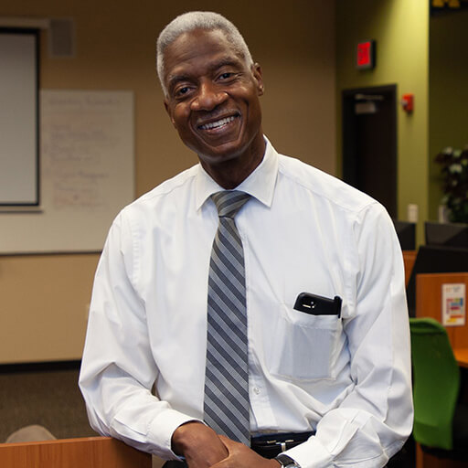 Dennis Bland, smiling in a long sleeved shirt and tie, leaning against a classroom desk.