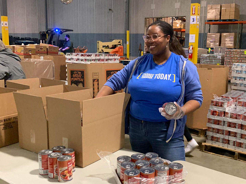 A worker in a food pantry warehouse with a big smile on her face packages canned food into cardboard boxes.