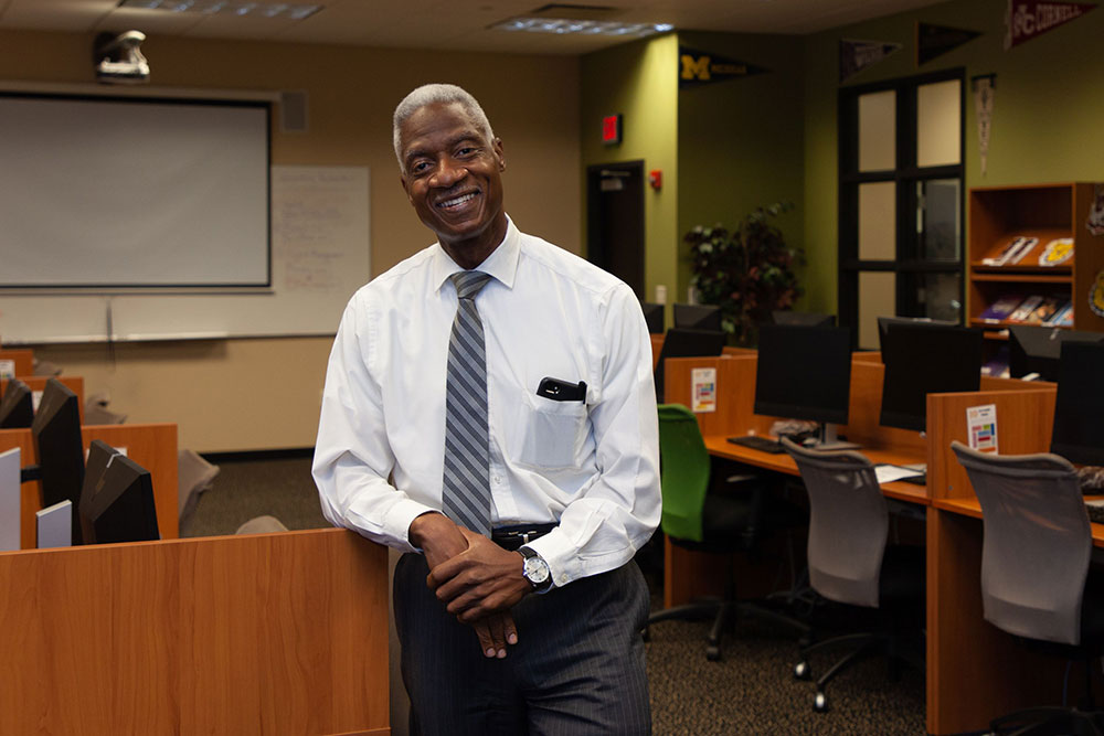 Dennis Bland, smiling in a long sleeved shirt and tie, leaning against a classroom desk.