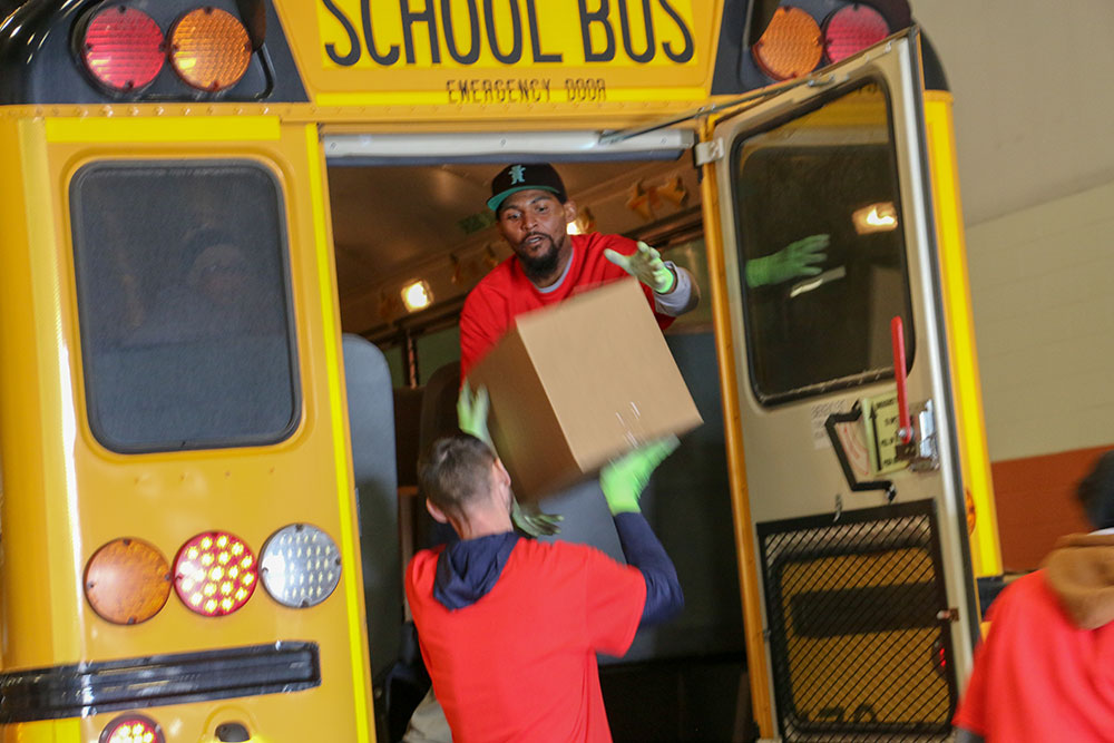 Worker on school bus hands box down to another worker on the ground from the back door of the bus.