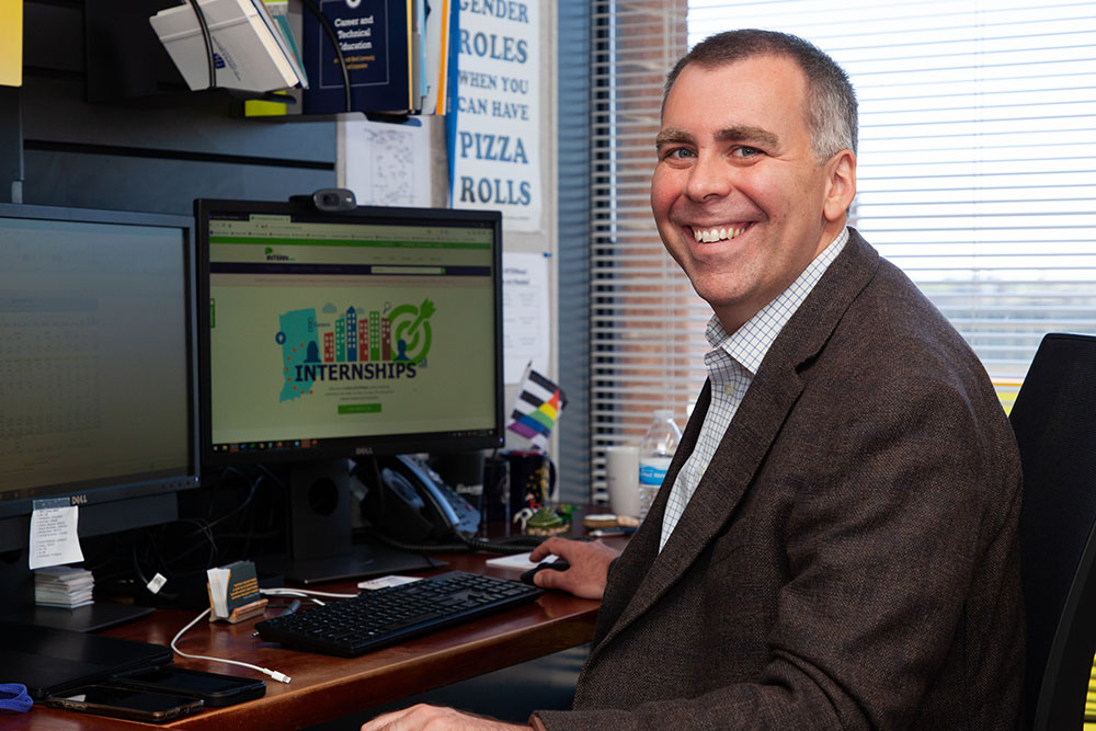 Mike Slocum of Indiana INTERNnet smiling at desk in an office high rise. A tablet, charter, and gay pride flag can be seen in his work environment.