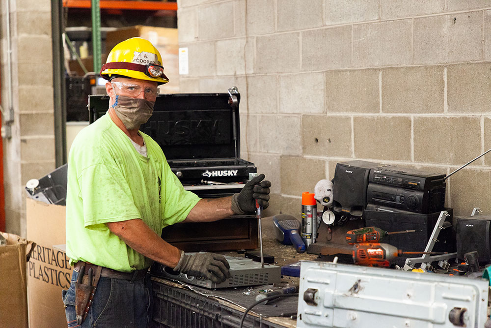 A man in a dirty fluorescent green shirt, plexi eye protection, and a yellow helmet with an attached work light is prying the plastic side off a video switcher with a long flat-head screwdriver. 