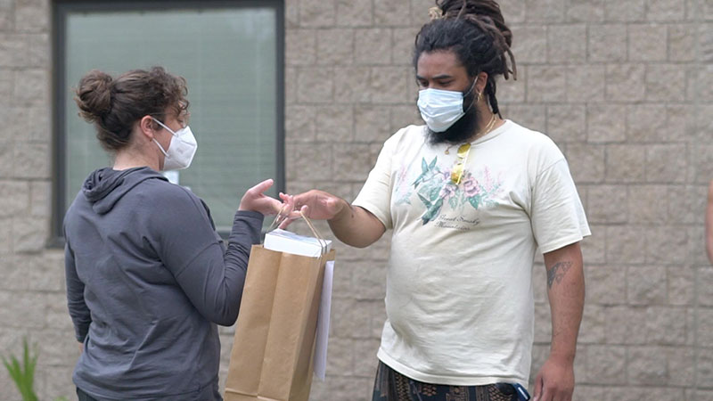 A food bank handoff, United Way employee hands over a bag to a customer, both are in COVID masks, outdoors.