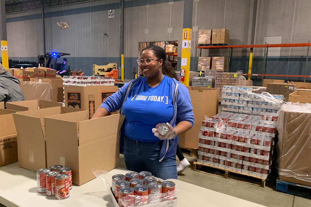 A worker in a food pantry warehouse with a big smile on her face packages canned food into cardboard boxes.