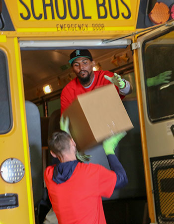 Worker on school bus hands box down to another worker on the ground from the back door of the bus.