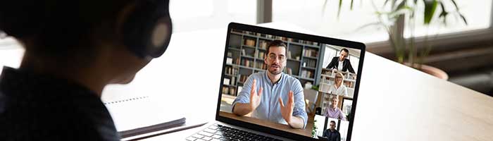 A man on a laptop screen delivers information to a Zoom chat. Four other people are seen onscreen. One person is offscreen taking notes and wearing headphones.