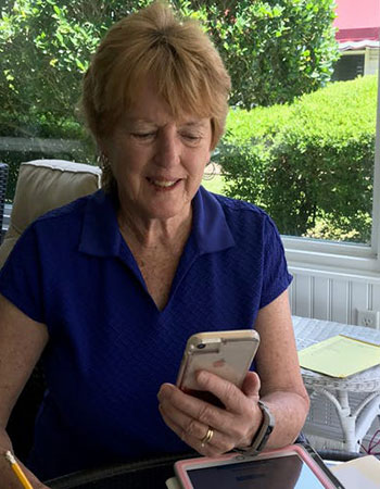 Nancy Gavin in blue shirt on back porch looking down at her mobile device, presumably doing mentoring work remotely. Green bushes fill out the background.