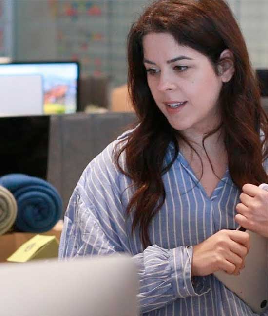 Brunette woman in vertically striped blue shirt clutches a closed laptop while listening to a seated colleague.