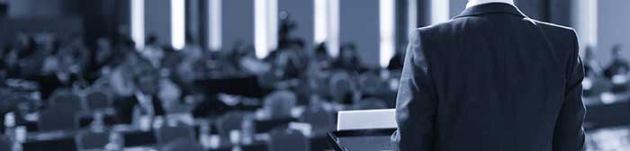 Faceless dude in a suit coat pictured from the back standing at a lectern addressing a blurry crowd of people.
