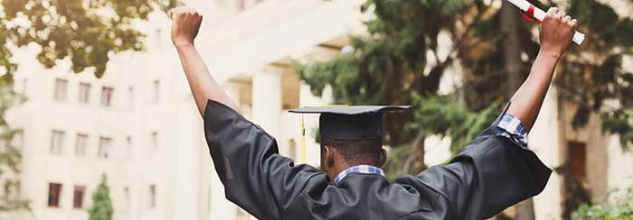 Black man at graduation holding his diploma victoriously high in the air.