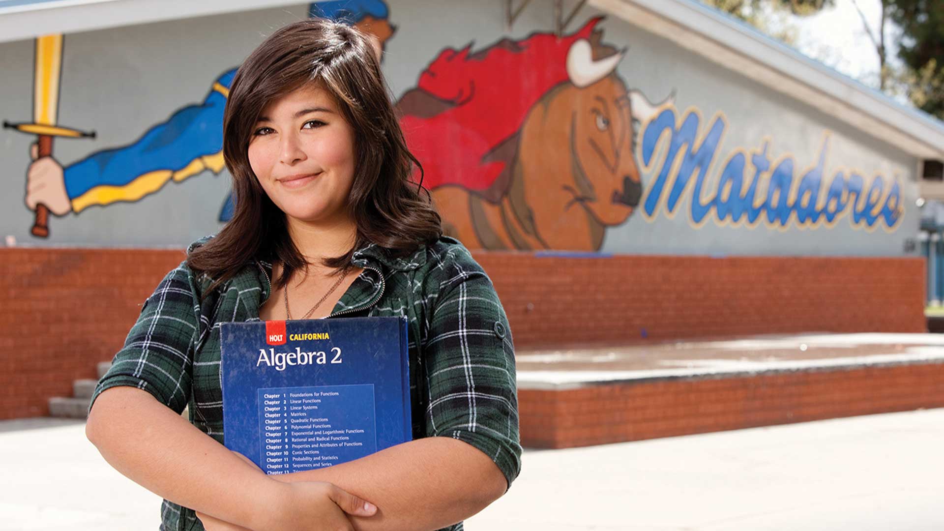 Latina student holding an algebra book standing in front of her school.