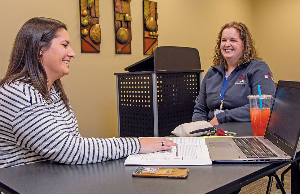 Two women casually working together and talking at a table with computer, cellphone, drinks, both women smiling.