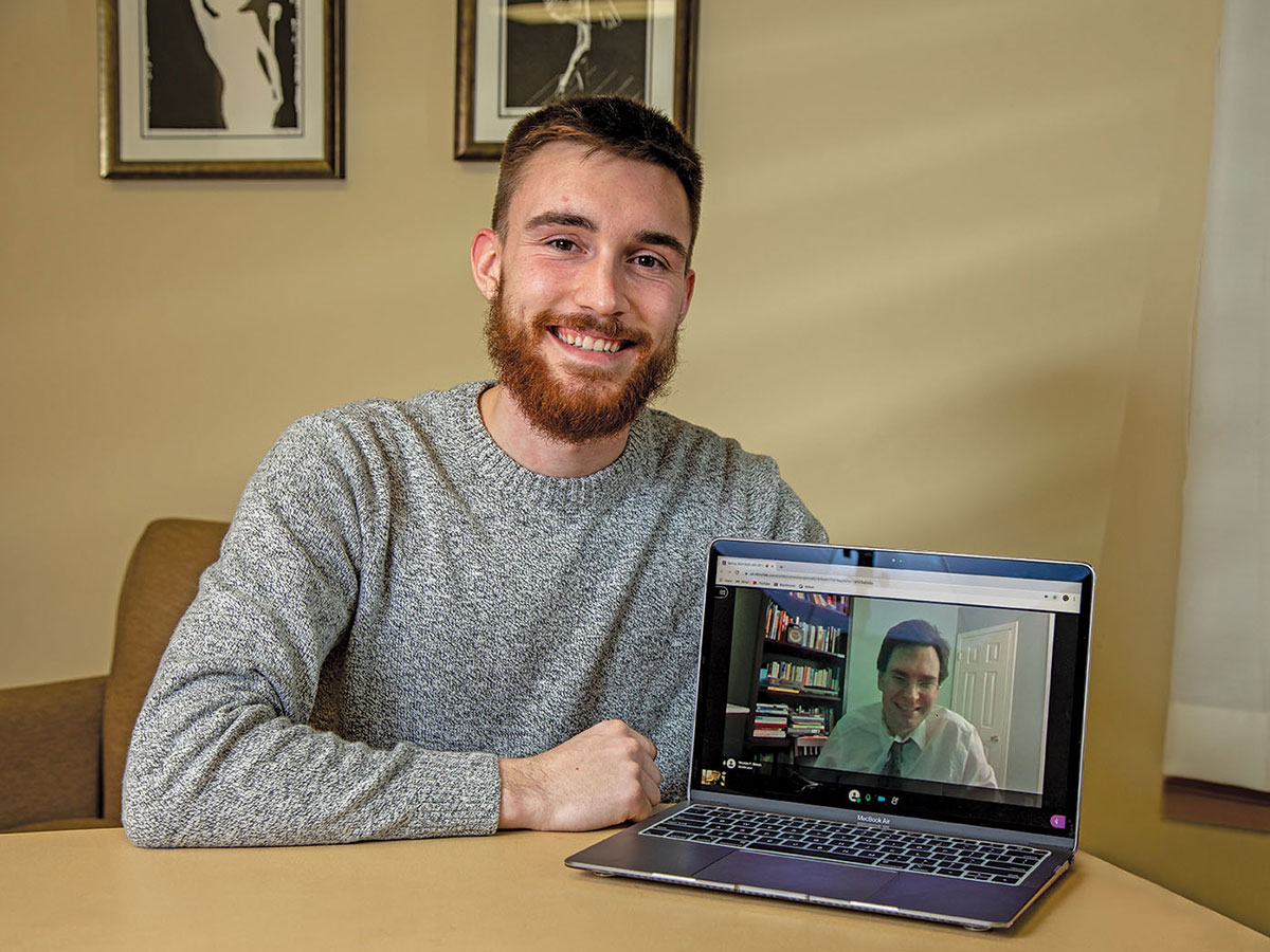 Brett Harrell smiling at a table. His laptop shows his a meeting attendee on screen.