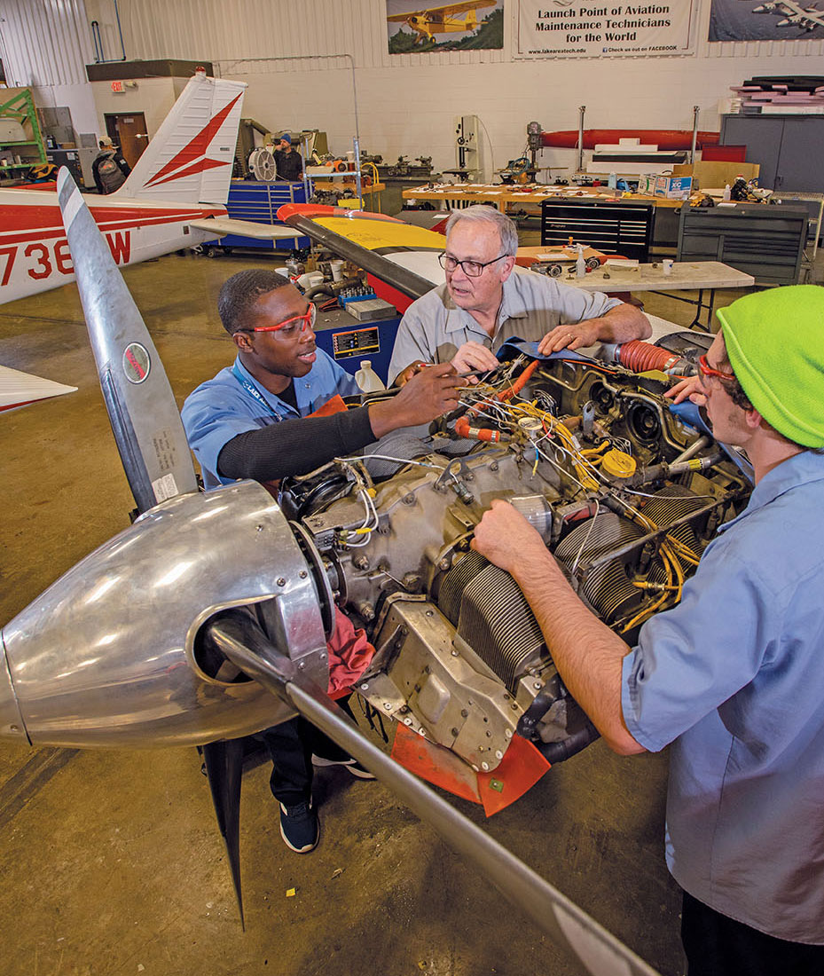 Three men work together in an airplane hangar. All have their forearms resting on an open airplane propeller engine.