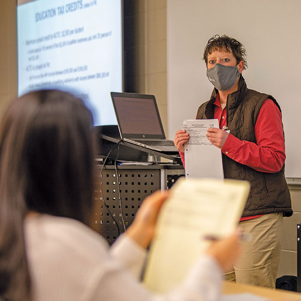 COVID-masked instructor in a vest leads a tax-preparation class. Her laptop is powerpoint is projected on a screen behind her.