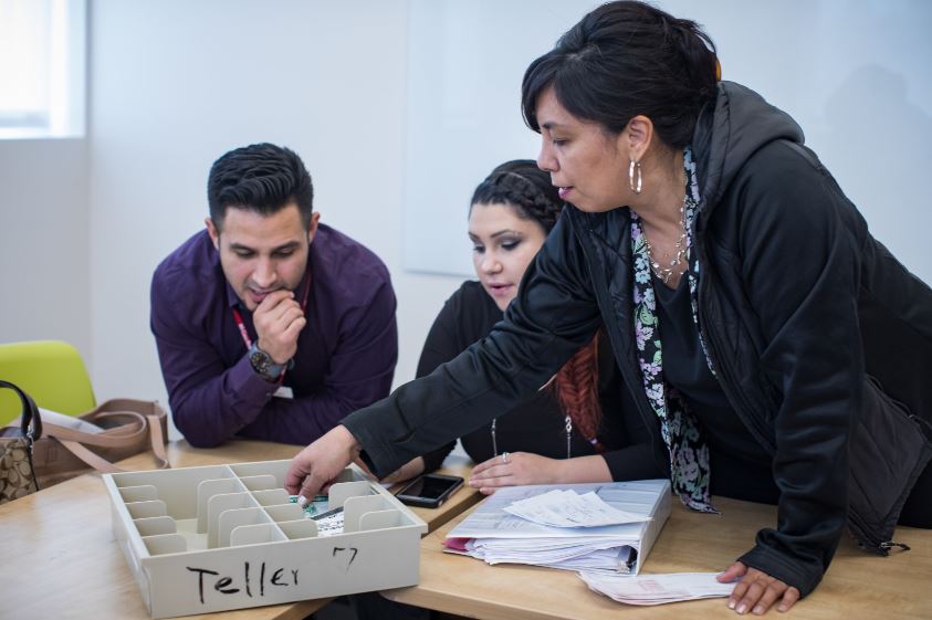 This photo shows a woman training two students to handle a cash drawer as they would at a bank.