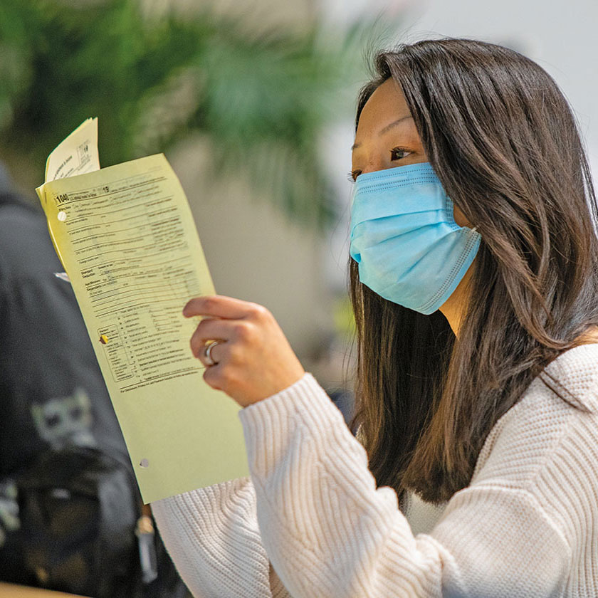 Same Asian woman from previous photo, here in a COVID mask, examines stapled sheets of a 1040 form.