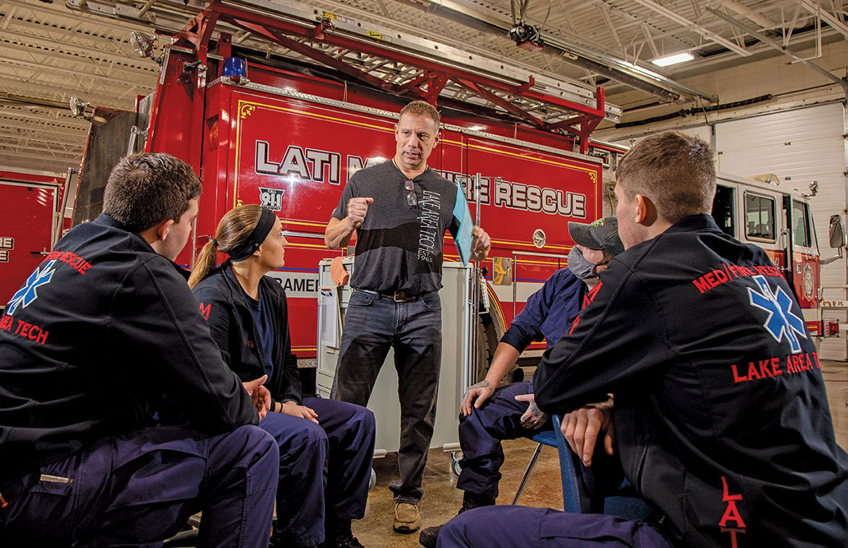 Seated students are instructed by a standing man. The students are wearing EMT uniforms. A fire truck in the background, the instruction takes place in an emergency medical garage.