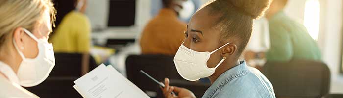 Black, female student in COVID mask talking with a fellow student, and holding a notepad.