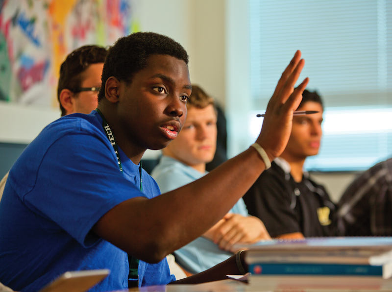 Young black man in a blue shirt, raising his hand in a classroom.