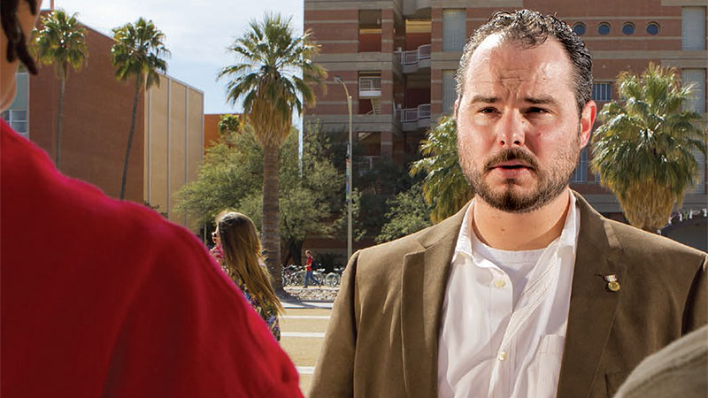 Rico Pereyda, outdoors in a brown suit coat, talking to unknown people. Palm trees and building in the background.