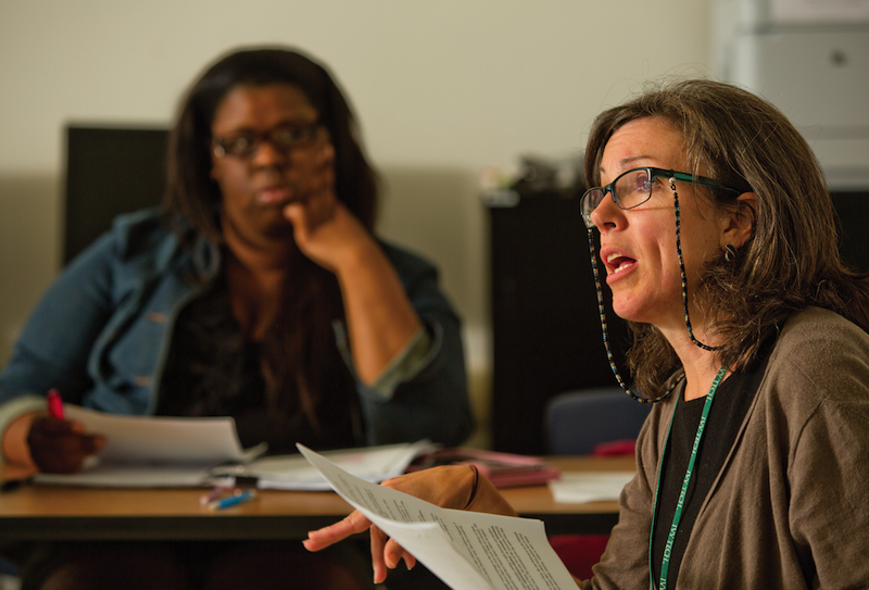 A seated woman holding a paper appears to be mid-sentence. Drooping from both sides of her glasses is a striped tether, like facial antennae.