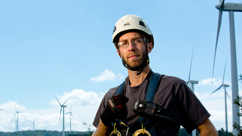 Wind turbine maintenance man Josiah Stanfill in full gear with wind turbines in the distance behind him.