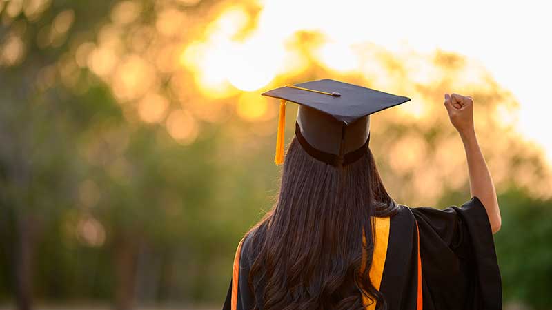 Woman in graduation mortar board raises her fist triumphantly toward the sunset.