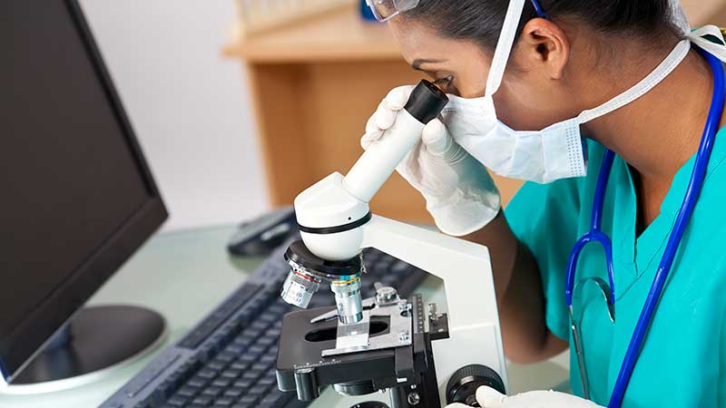 COVID-masked woman peers into a microscope.