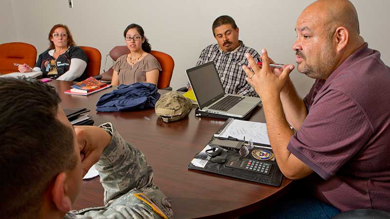 Navy veteran John Vasquez sits at the head of an oval table talking to a group of veterans.