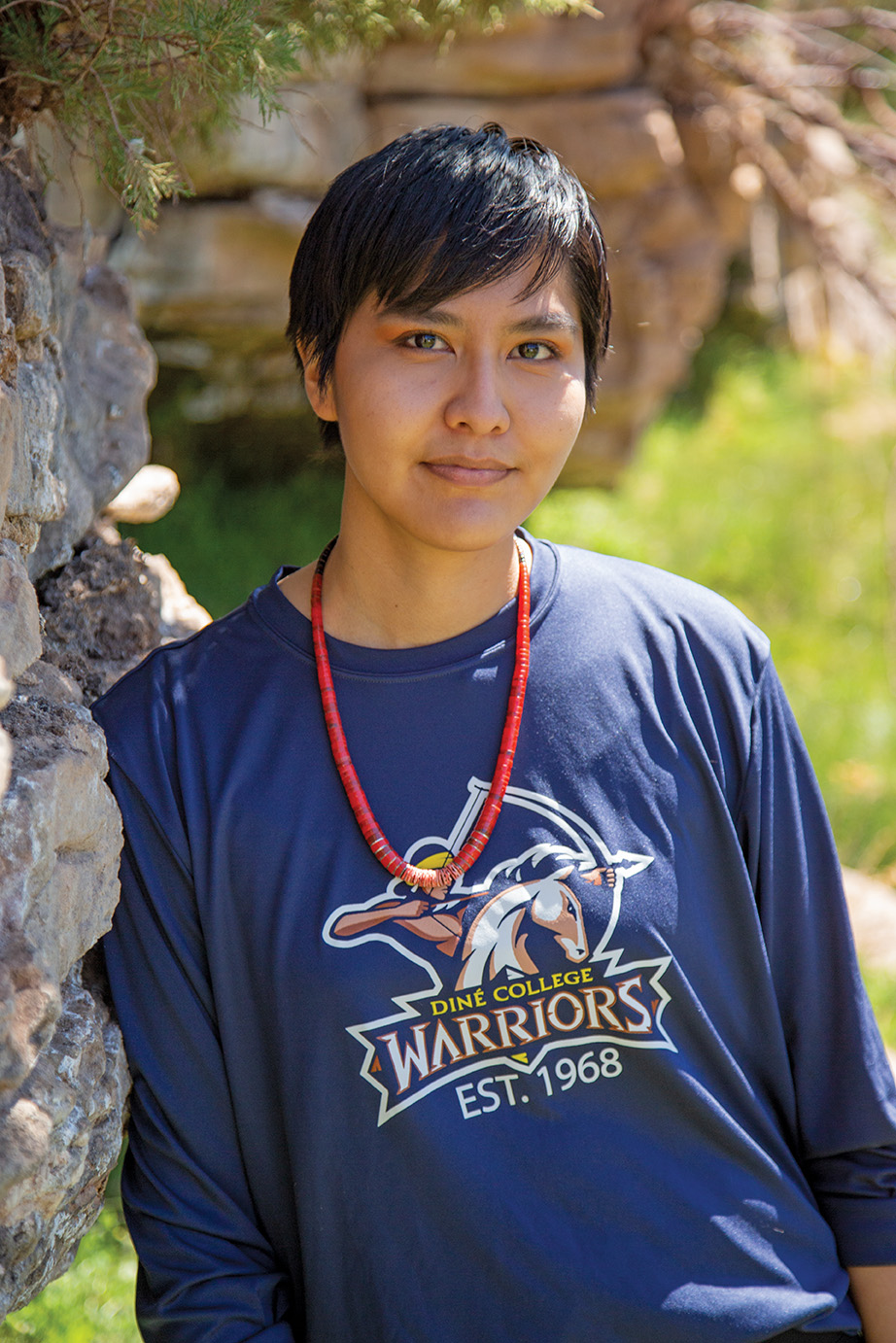 Female Navajo student in Diné College Warriors sweatshirt leans agains the rocks.