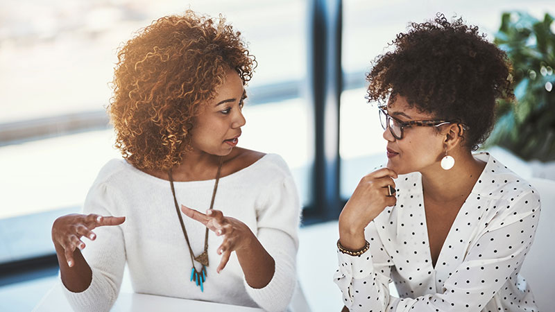 Two women of color strategize and converse at a desk.