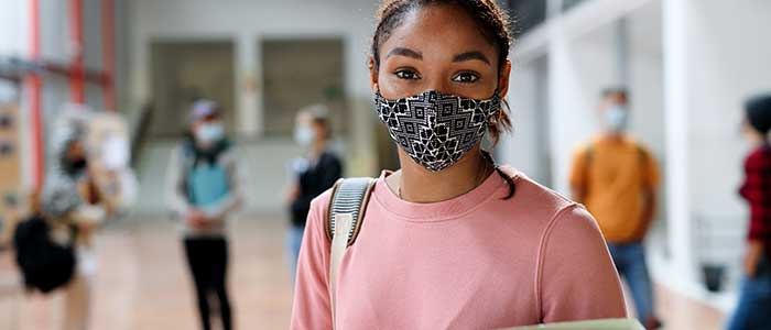 Black, female student in a pink sweater and covid mask, holding a pastel green binder. Other students can be seen chatting in the background.