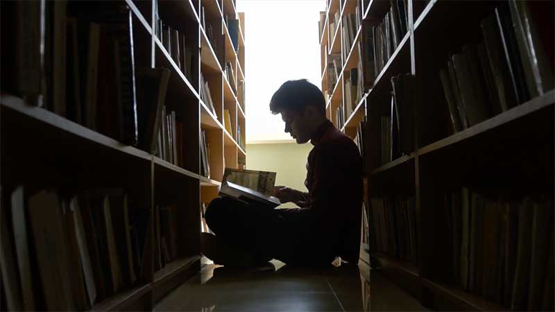 Man in library stacks in profile, sits reading.