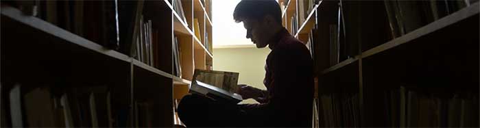 Man in library stacks in profile, sits reading.