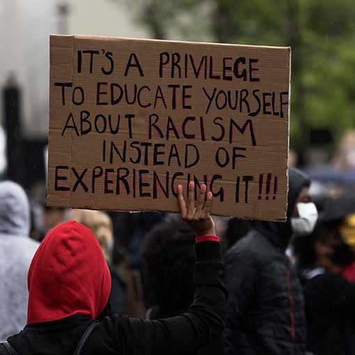 In a faceless crowd, a sign is held up. It reads, “It's a privilege to educate yourself about racism instead of experiencing it!!.”