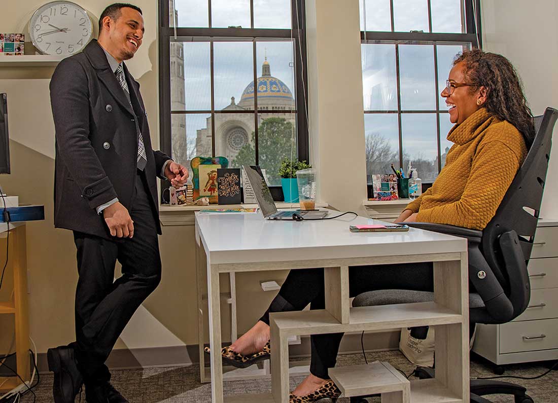 Ariel Ventura-Lazo standing in front of the desk of Nicole Lynn Lewis. They are smiling and apparently conversing.