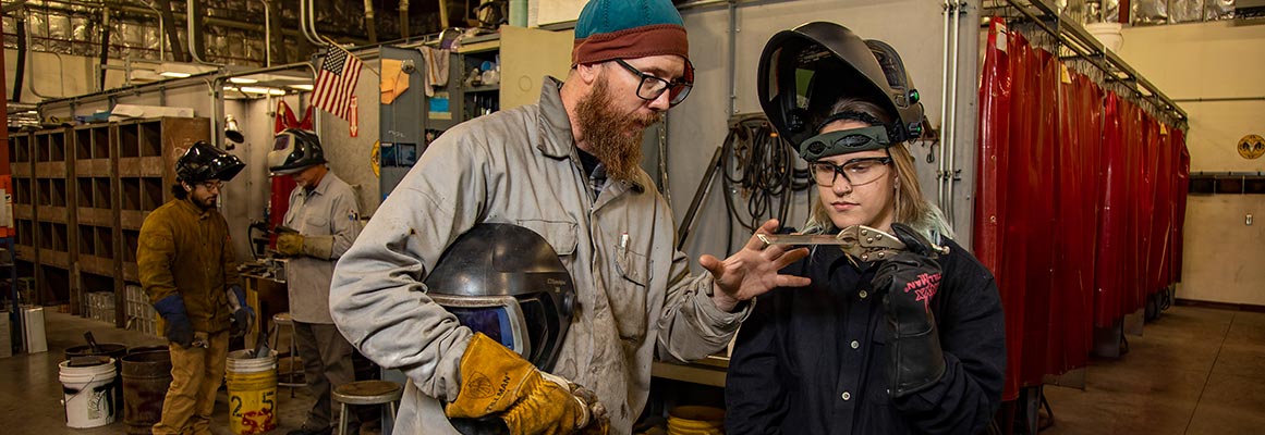 Cat, the welder featured in the Spring 2023 issue of Focus magazine is working with a colleague. She's gripping a small sheet of metal with vice grips.
